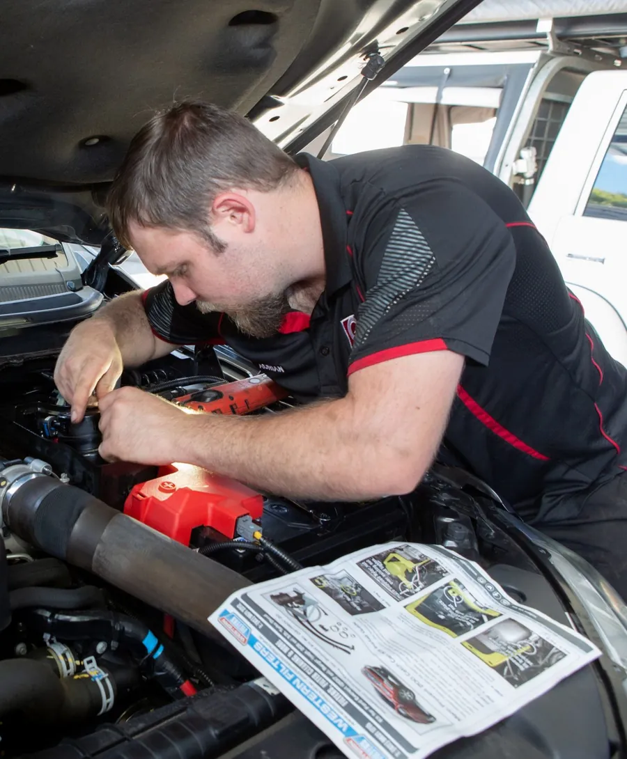 A mechanic working in an engine bay with the service manual open beside him
